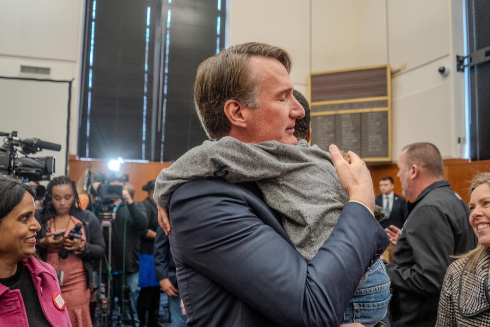 Glenn Youngkin hugs a young supporter in a crowded room as press cameras look on.