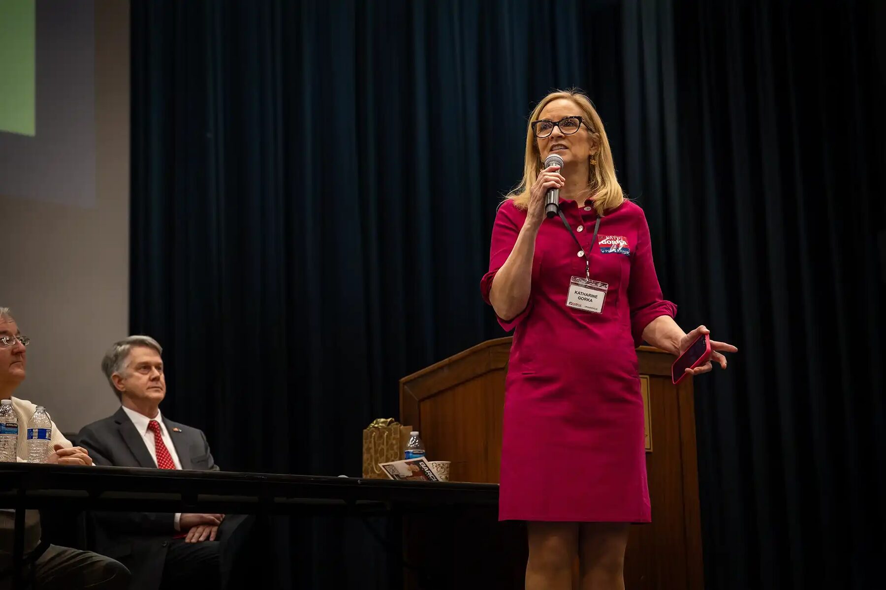 Chairwoman Katie Gorka, in a magenta dress with her convention name badge, addresses committee members with a handheld microphone from a dark stage, two officials seated at a table behind her.